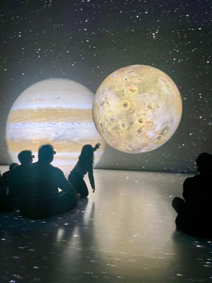 A projected Jupiter and its moon Io in the AELINZ exhibition, with visitors seated on the floor