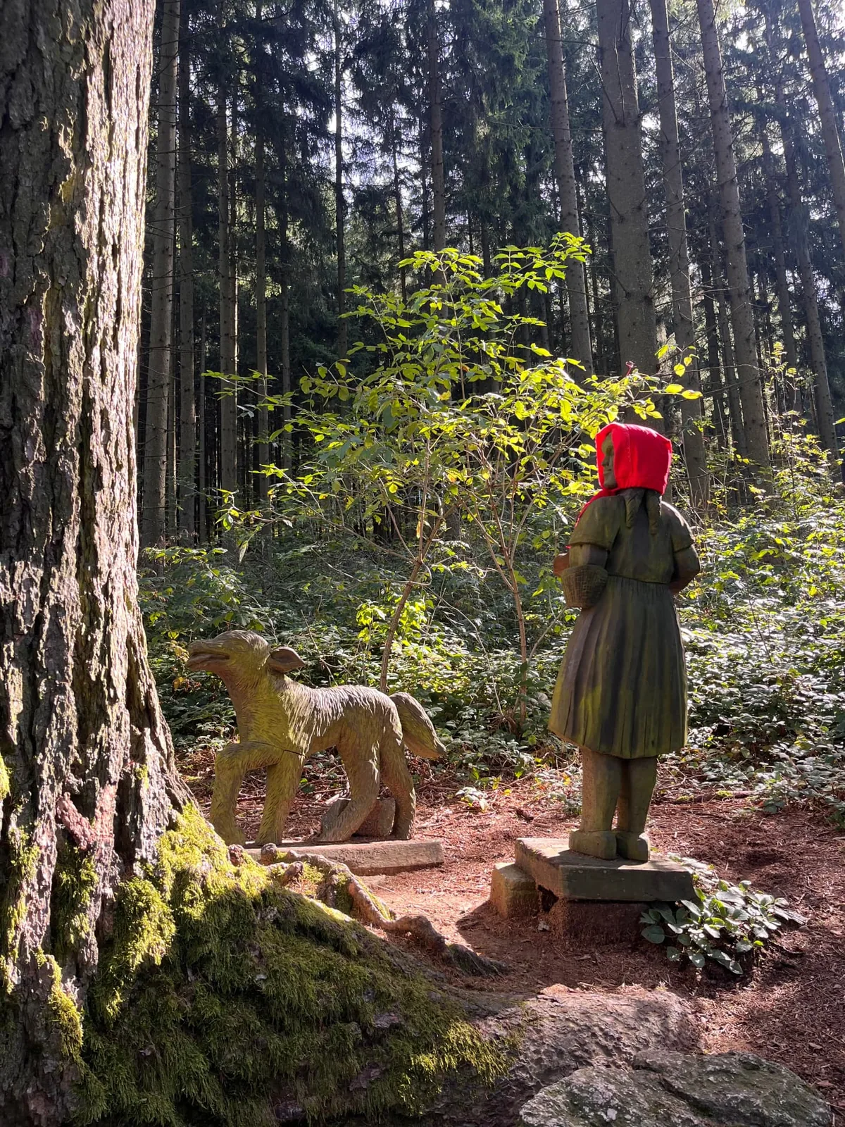 Carved wooden figures from the Little Red Riding Hood fairy tale along a sunlit forest path